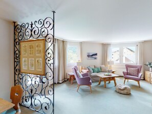 Ein helles Wohnzimmer mit einer Couch, zwei Sesseln, einem Couchtisch und einer dekorativen Metalltrennwand mit gerahmten Dokumenten.