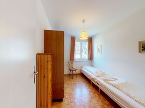 Una habitación pequeña y bien iluminada con un armario de madera, una cama individual con ropa de cama blanca, una silla y una ventana con cortinas.