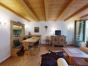 Ein gemütliches Zimmer mit Holzbalkendecke, einem Esstisch mit Stühlen, einem Fernseher und einem Kuhfellteppich.