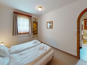 A bedroom with two beds, a window with curtains, a radiator, a painting, and an arched doorway leading to another room. - Apartment Larschigna - Attraktive 3.5-Zimmerwohnung by Interhome (Zuoz)