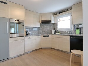 A modern kitchen with a built-in oven, microwave, and dishwasher, light-colored cabinets, granite countertops, and a window providing natural light. - Apartment Falcun by Interhome (Celerina)