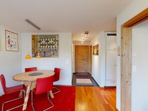 Ein Essbereich mit einem runden Holztisch und roten Stühlen, ein Flur mit einer Holztür sowie eine Küche mit weißen Schränken.
