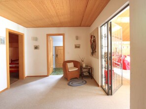 Ein Zimmer mit Holzbalkendecke, einer Tür zu einem anderen Raum, einem Sessel, einem kleinen Tisch mit einer Vase und einer Glastür mit Blick.
