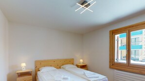 Une chambre à coucher avec un lit en bois, deux tables de chevet avec des lampes, une fenêtre avec des stores et un radiateur.