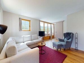 A modern living room with a sofa, armchair, and ottoman, a television, and a large window with a view of a balcony. - Apartment Martina by Interhome (Pontresina)