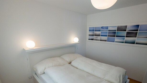 Ein Schlafzimmer mit einem Bett, einer Kopfteil, einem an der Wand befestigten Regal mit einer Lampe und einer Reihe von Fotos.