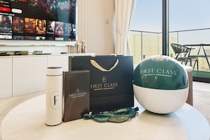 A table with a First Class branded water bottle, a thermos, a bag, and a pillow on a balcony with a TV displaying movie posters in the background. - FIRST CLASS |1BR| Beachfront with Ain Dubai Views (Dubai Harbour)