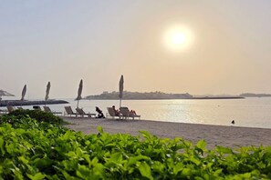 A beach with sun umbrellas and lounge chairs, a calm sea, and a distant shoreline. - FIRST CLASS |1BR| Beachfront with Ain Dubai Views (Dubai Harbour)