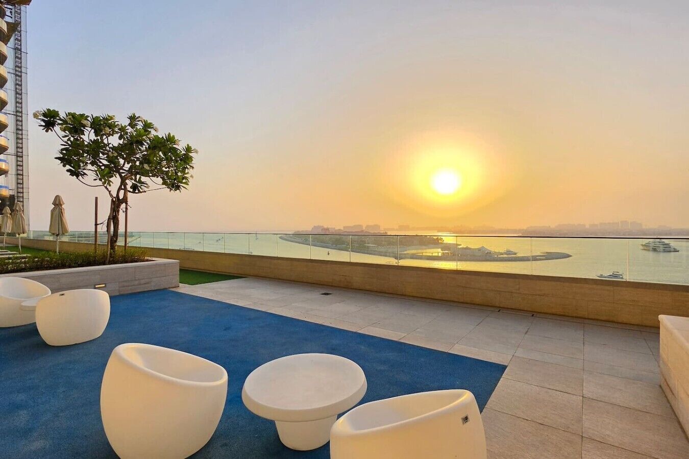 Une terrasse sur le toit offrant une vue sur le coucher de soleil sur l’eau, avec des sièges modernes et un petit arbre.