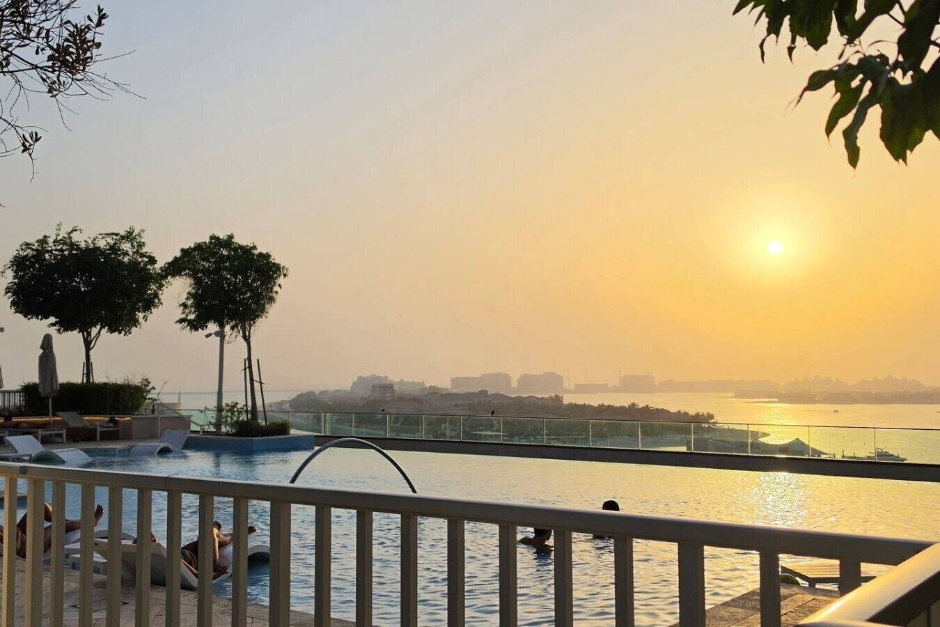 Un espace piscine avec des chaises longues et une vue sur le coucher de soleil au-dessus d’une étendue d’eau.