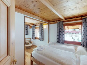A hotel room with a wooden ceiling, two beds, a chair, and a view of a building outside. - Apartment Stalla Bodmer by Interhome (Samedan)