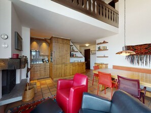 A spacious living area with a dining table, red and blue chairs, a fireplace, and a kitchen in the background. - Apartment Gianda Verda C31 by Interhome (Silvaplana-Surlej)