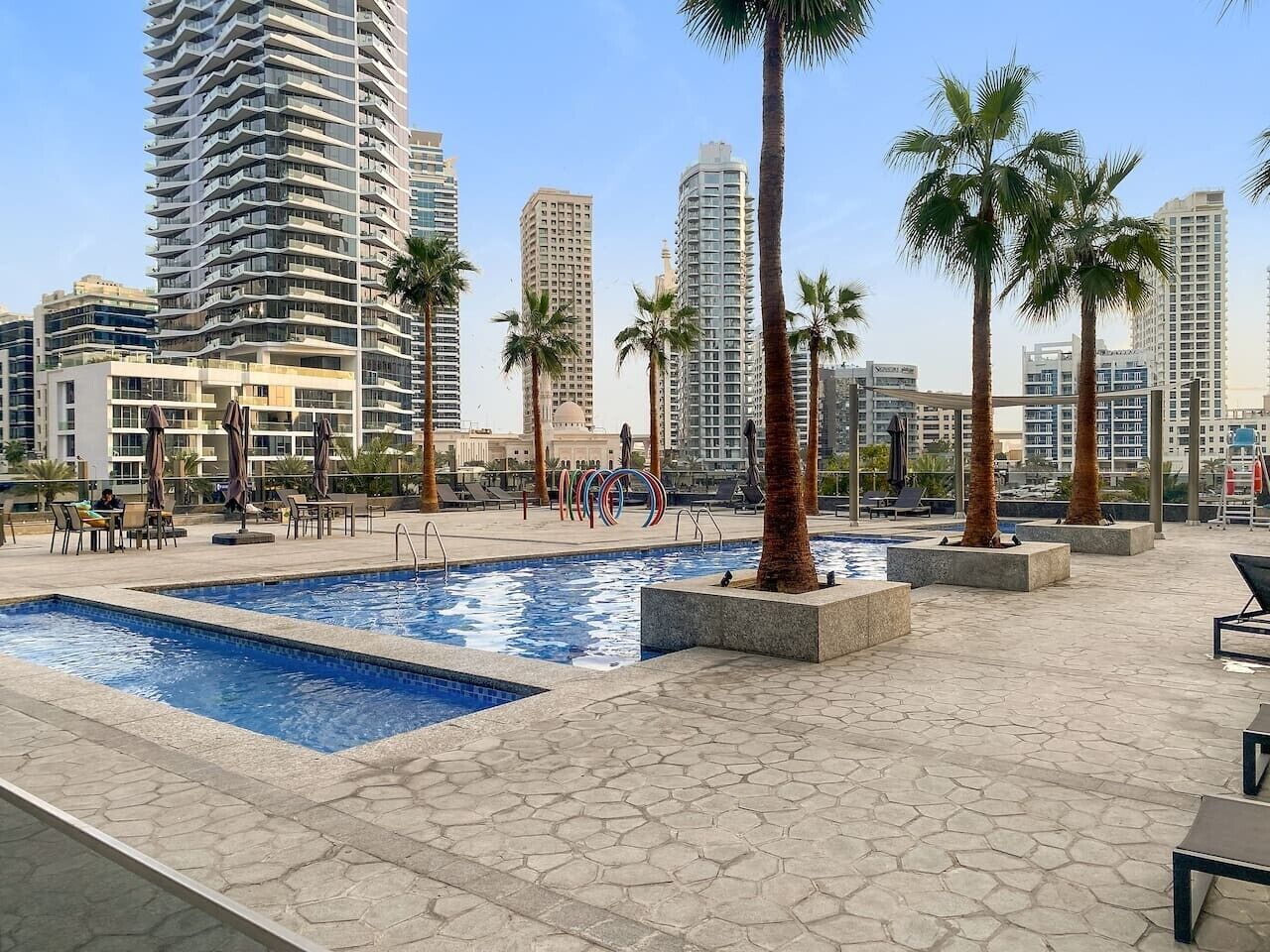 A rooftop pool area with palm trees, lounge chairs, and a view of surrounding high-rise buildings.