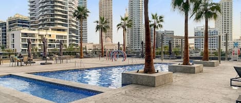 A rooftop pool area with palm trees, lounge chairs, and a view of surrounding high-rise buildings.