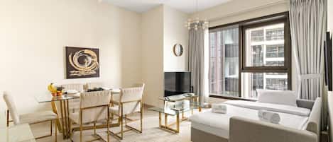 A modern living room with a glass dining table, comfortable seating, a flat-screen TV, and a large window with curtains.