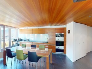 A modern kitchen with wooden cabinets, a dining area with a glass table, and a view through large windows. - Apartment Quadratscha by Interhome (Samedan)