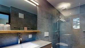 Un baño moderno con un espejo grande, un tocador de madera, un lavamanos blanco y una zona de ducha con mampara de vidrio.
