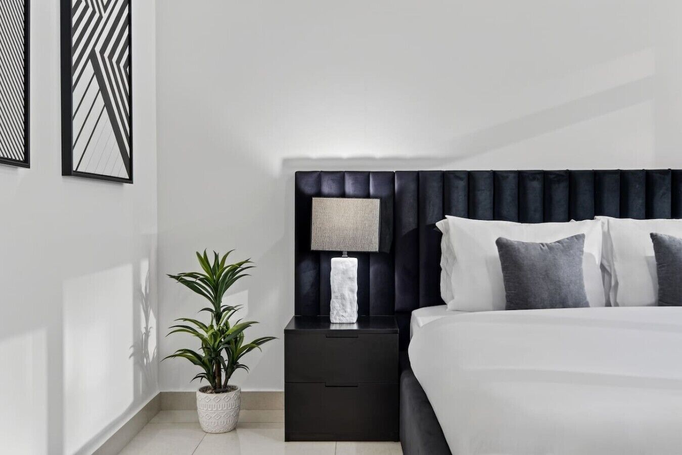 Un dormitorio moderno con cabecera oscura, una mesita de noche con lámpara y una planta en maceta.