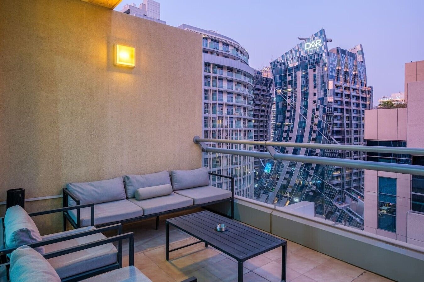 A balcony with a sofa and a table, overlooking a cityscape with modern buildings.