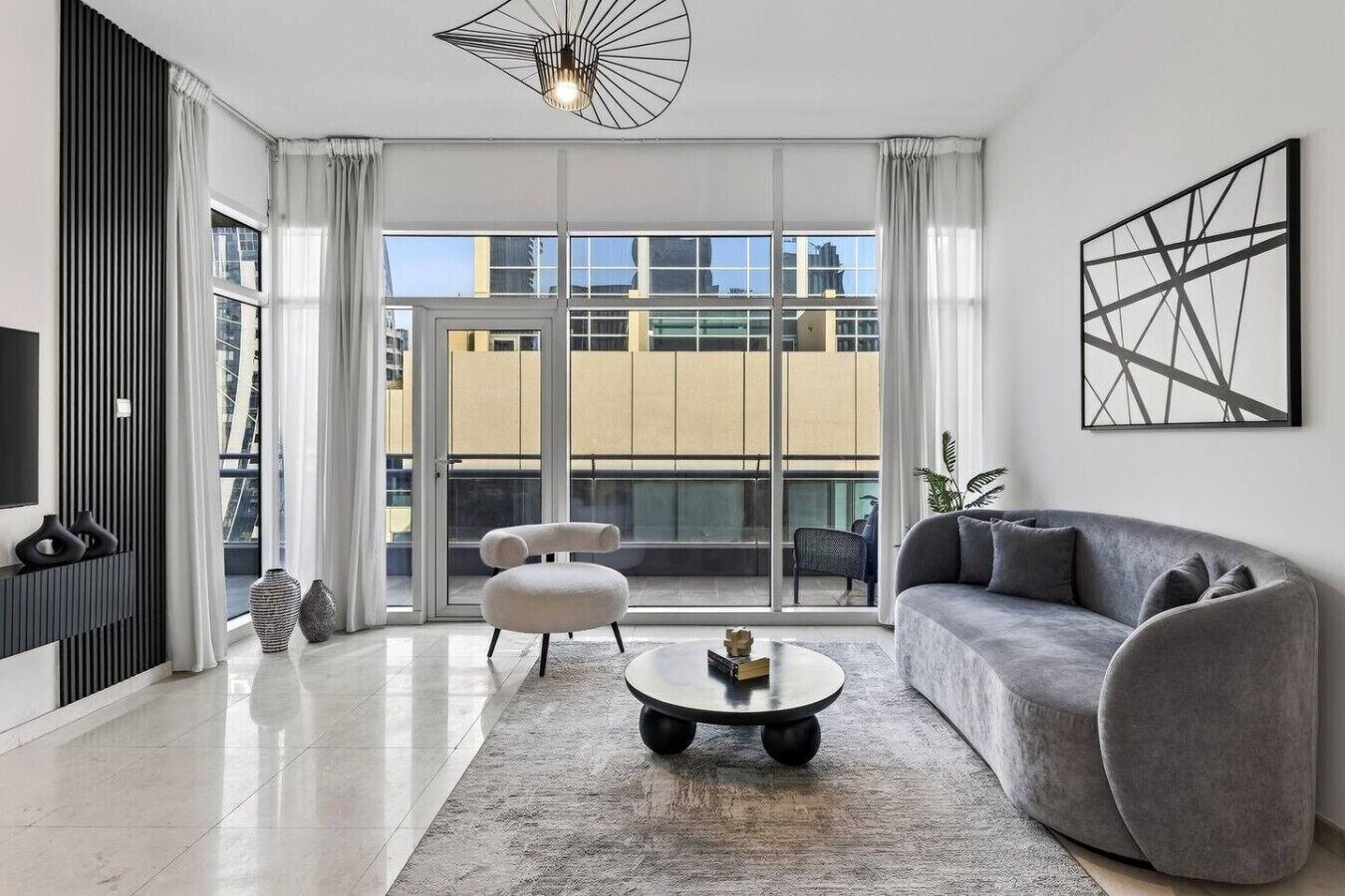 Ein modernes Wohnzimmer mit einer Couch, einem Couchtisch und einem Sessel. Ein großes Fenster mit Vorhängen bietet Blick auf Gebäude, und eine Deckenleuchte sorgt für Licht.