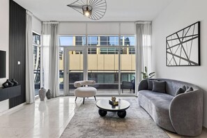 A modern living room with a sofa, a coffee table, and a chair. There is a large window with curtains, a view of buildings outside, and a ceiling light fixture. - FIRST CLASS | 2BR | Luxury Retreat | Near Downtown (Business Bay)