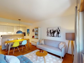 Ein modernes Wohnzimmer mit Sofa, einem Esstisch mit Stühlen und einer Küchenzeile im Hintergrund.