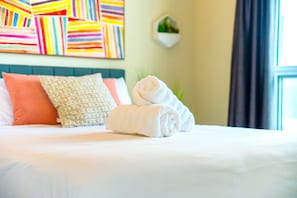 Un lit bien fait, avec une serviette enroulée, un coussin à motifs et une œuvre d’art colorée à rayures accrochée au mur.