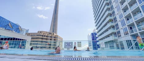 A swimming pool with people, a tall tower, and modern buildings.