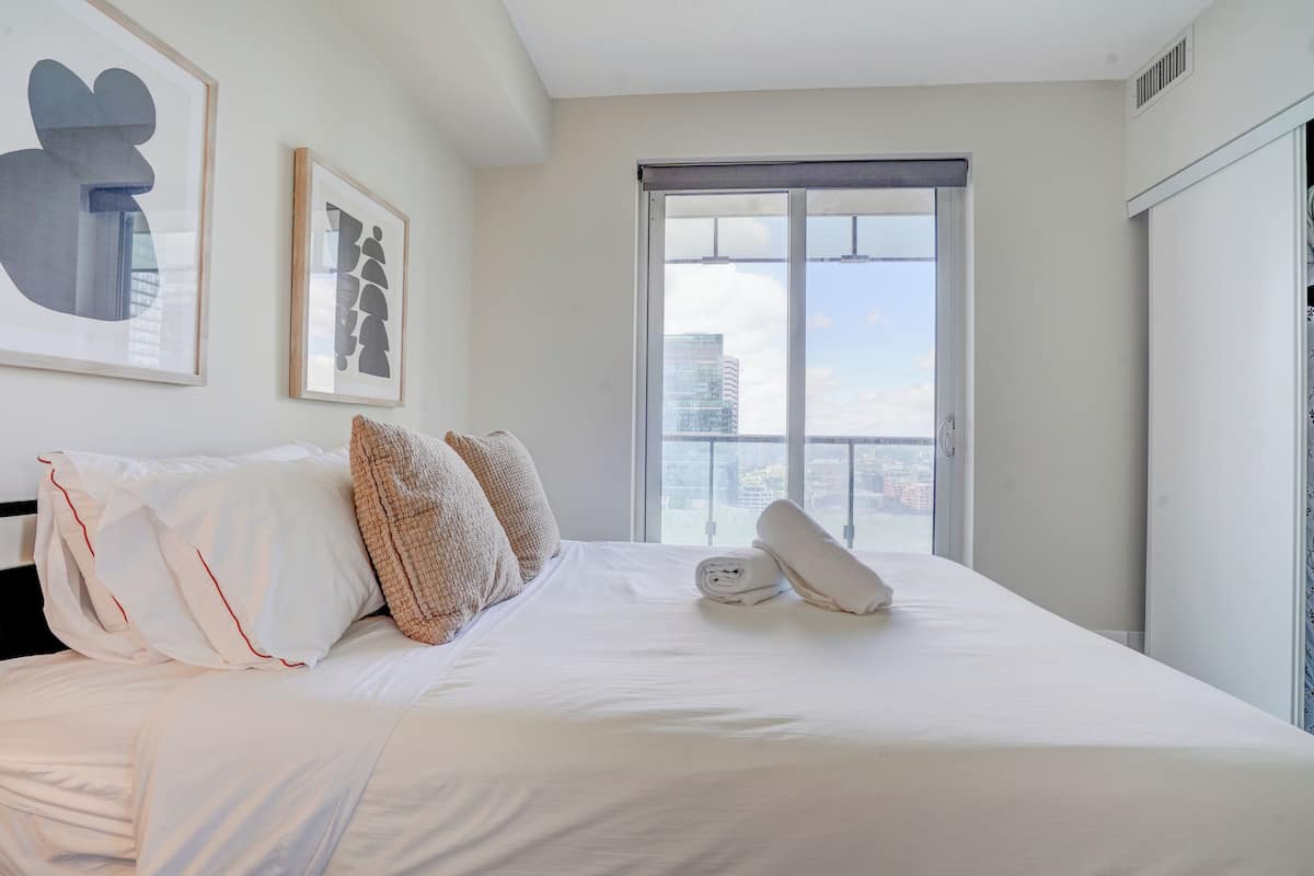 Habitación de hotel con una cama grande, dos almohadas, una ventana con vista a la ciudad y cuadros en la pared.