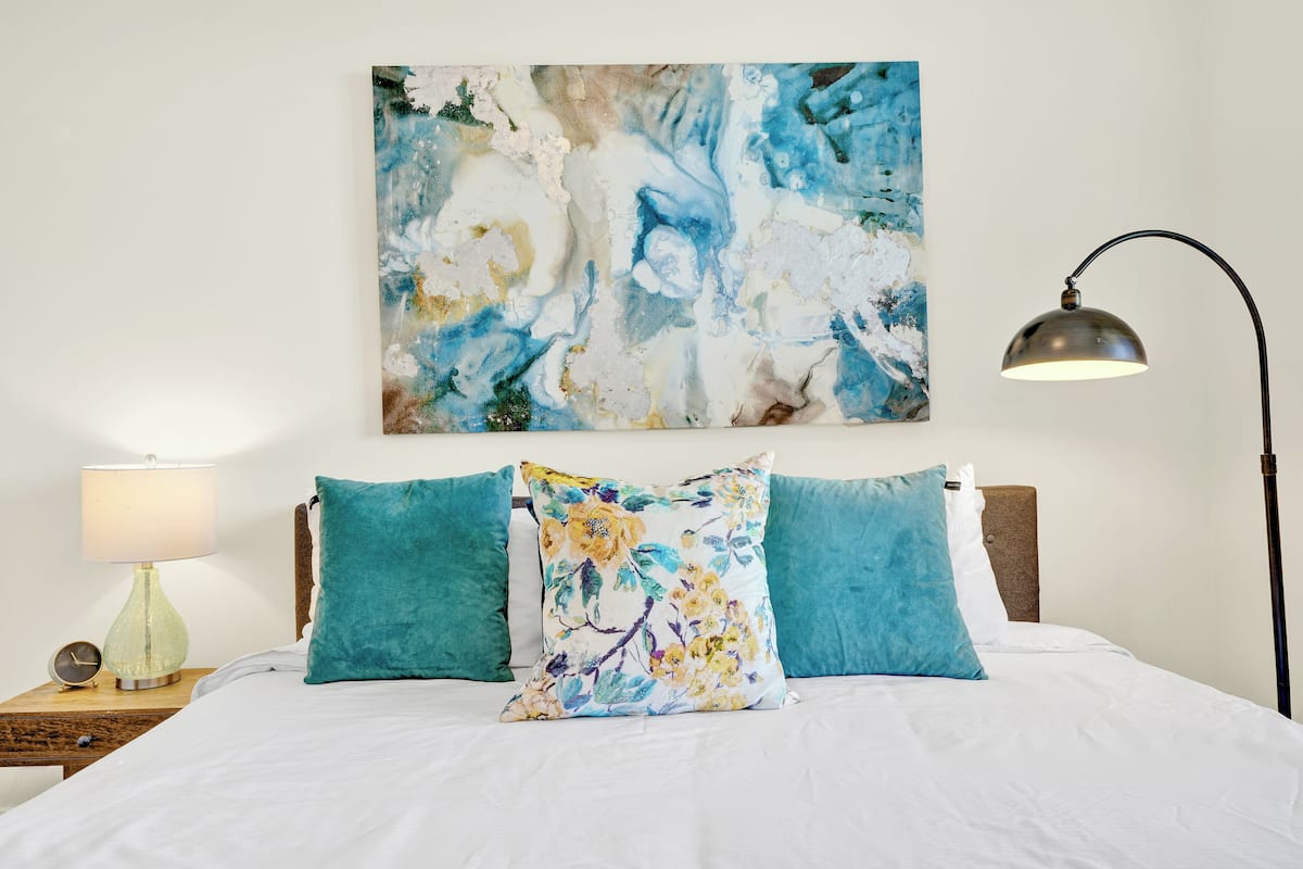 Una cama bien hecha con un cojín estampado y un cuadro abstracto colgado en la pared, además de una lámpara de noche.