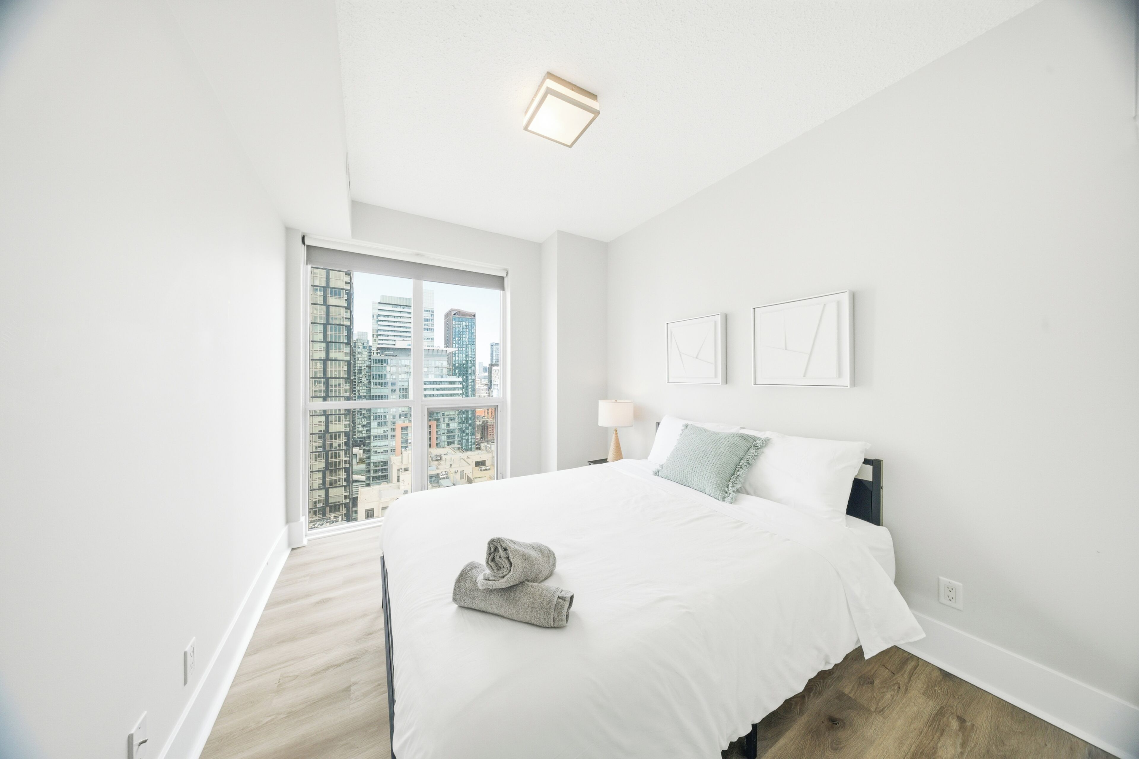 A modern bedroom with a large bed, a view of the city through a floor-to-ceiling window, and a small bedside table with a lamp.