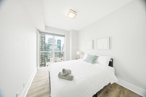 Un dormitorio moderno con una cama grande, vistas de la ciudad a través de un ventanal de pared a techo, y una mesita de noche pequeña con una lámpara.