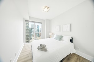 A modern bedroom with a large bed, a view of the city through a floor-to-ceiling window, and a small bedside table with a lamp.