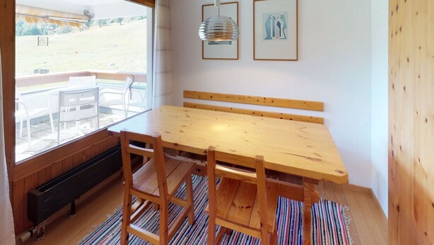 Une table à manger en bois, avec des chaises, un banc et une vue sur un espace extérieur verdoyant, visible à travers de grandes fenêtres.