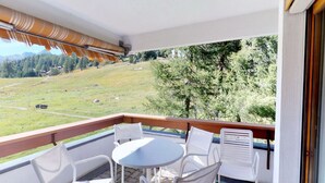 Un balcon avec une table et des chaises, donnant sur un paysage verdoyant parsemé d’arbres et surmonté, au loin, d’une montagne.