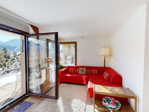 Ein modernes Wohnzimmer mit einem roten Sofa, einem kleinen Tisch und einer Lampe. Eine große Glasschiebetür führt auf einen Balkon mit Blick auf schneebedeckte Bäume und Berge.