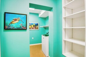 Ein Zimmer mit türkisfarbenen Wänden, einem Bild einer gerahmten Meeresschildkröte, einer Waschmaschine und leeren Regalen.