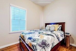 Een slaapkamer met een bed, nachtkastje, raam met jaloezieën en een houten vloer.