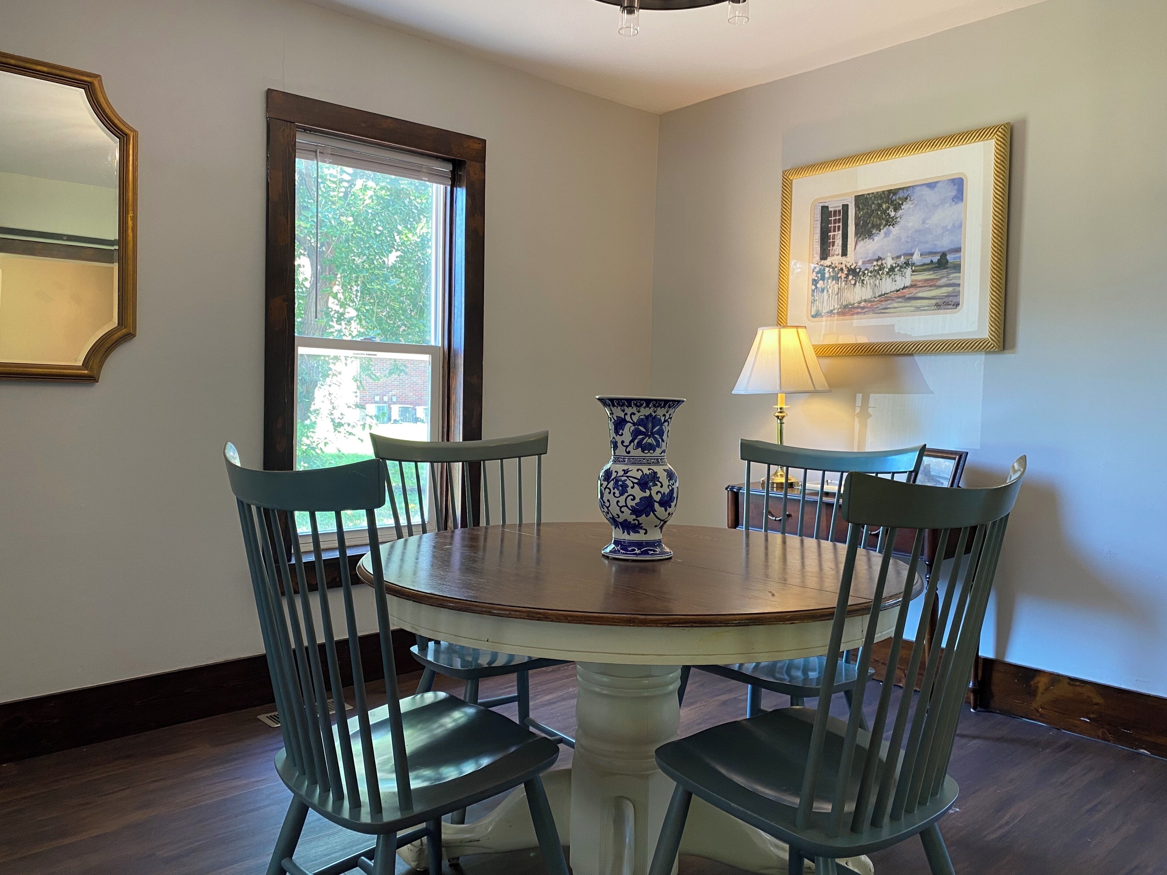 A dining area with a round table, four chairs, a vase, a lamp, and a framed picture on the wall.
