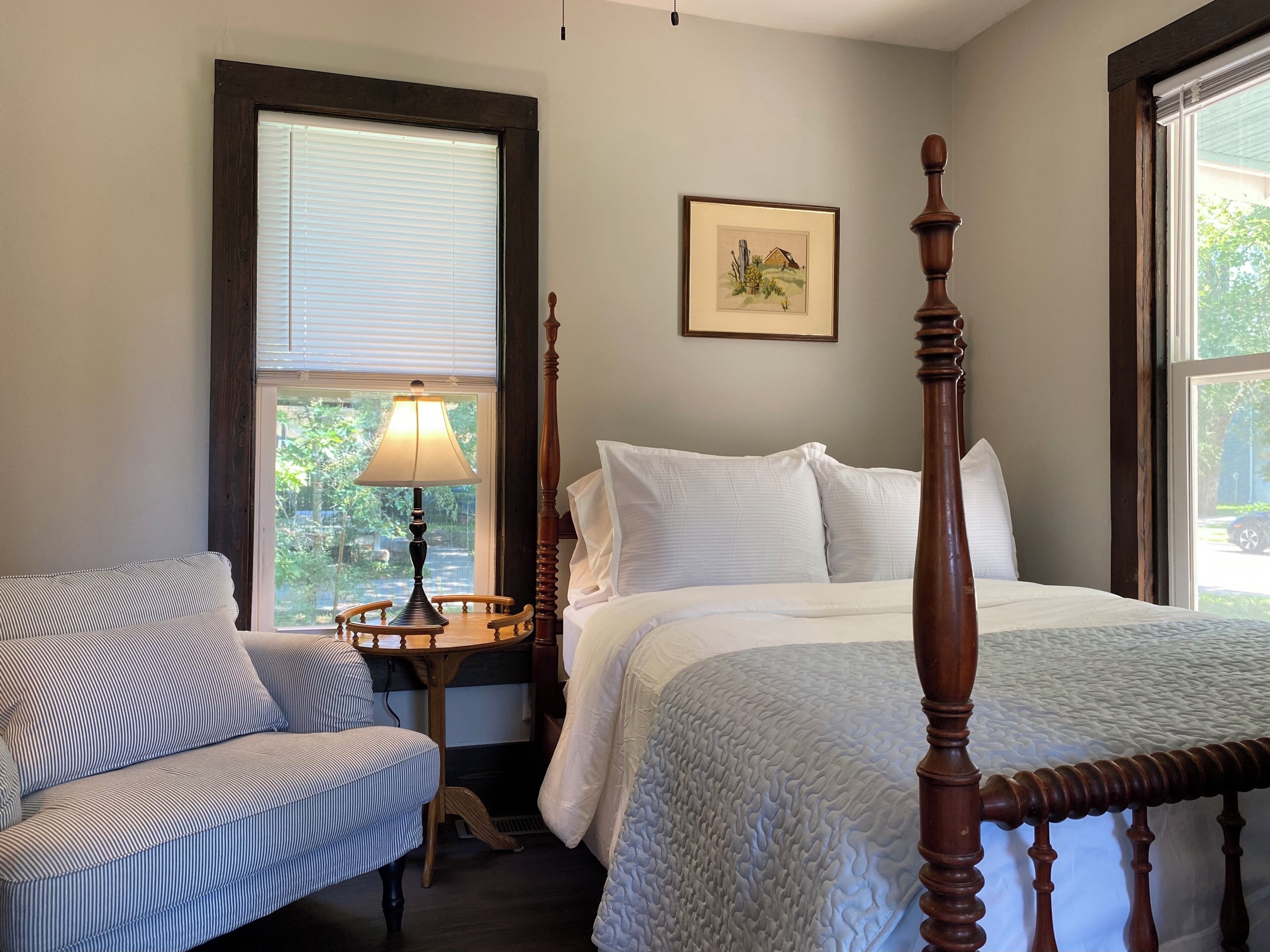 A bedroom with a four-poster bed, a bedside table with a lamp, a chair, and a framed picture on the wall.