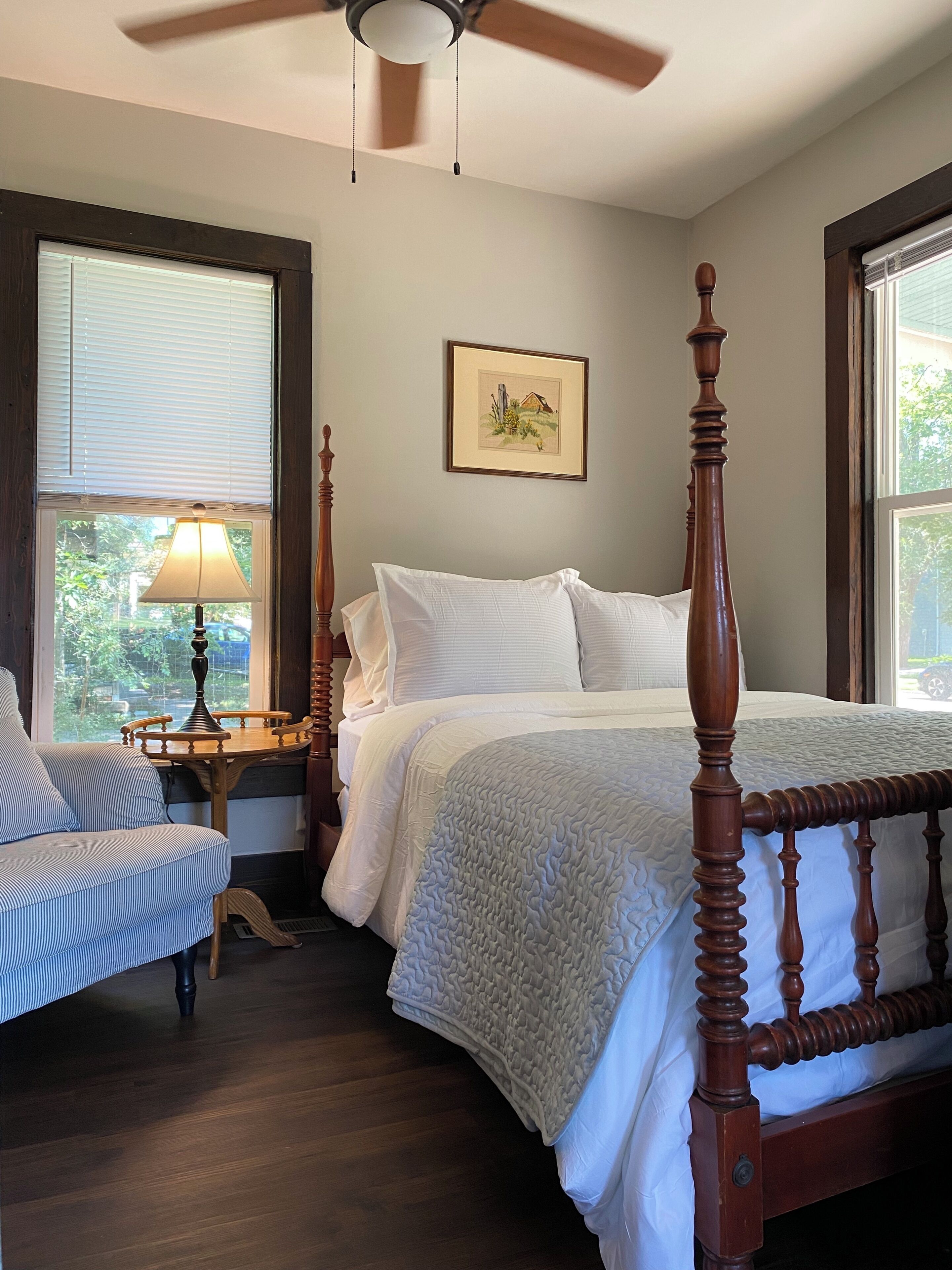 A bedroom with a four-poster bed, a bedside table with a lamp, a chair, and a framed picture on the wall.