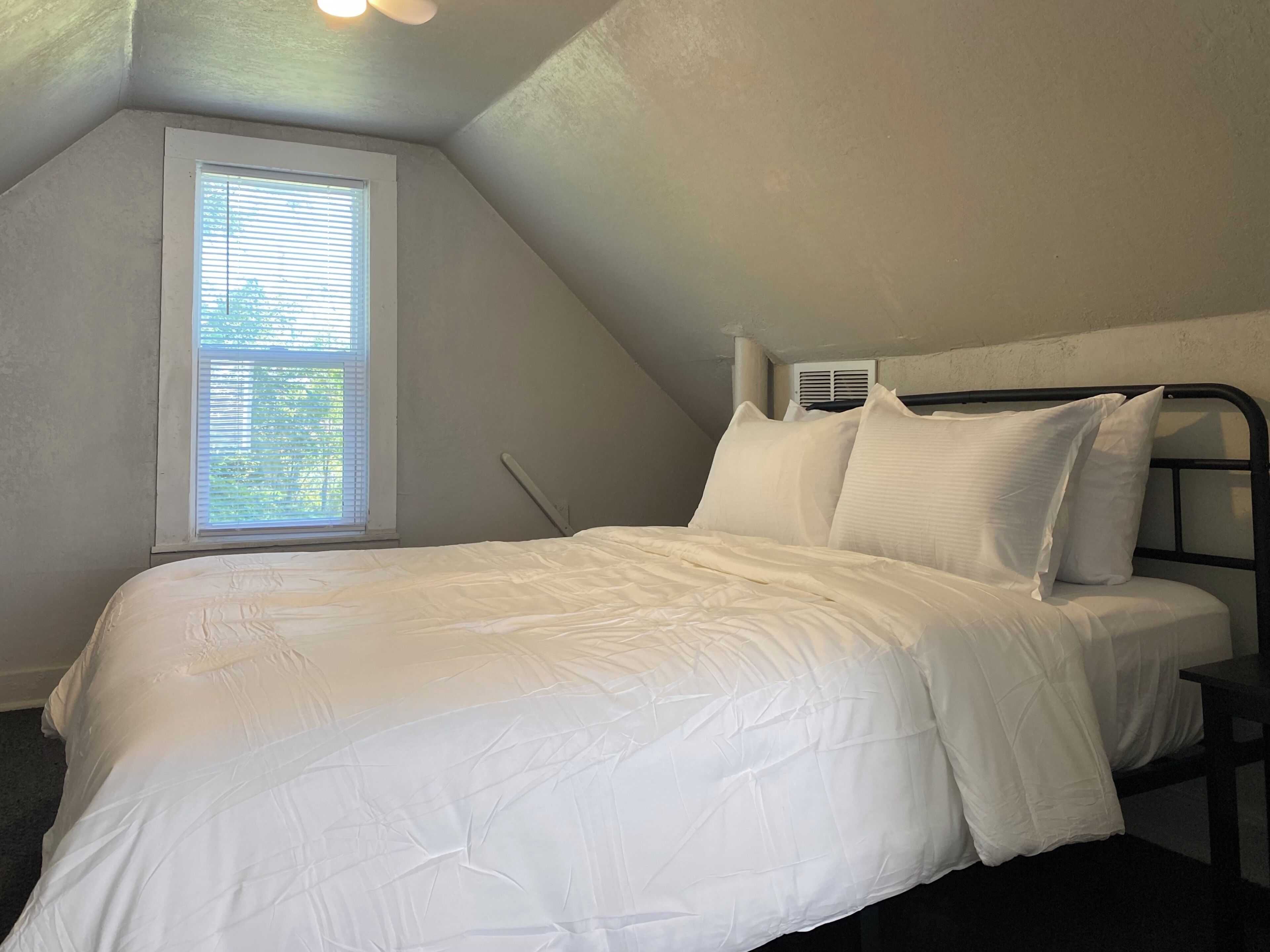 Une chambre à coucher avec un lit, du linge de lit blanc, une fenêtre avec des stores et un appareil de climatisation.