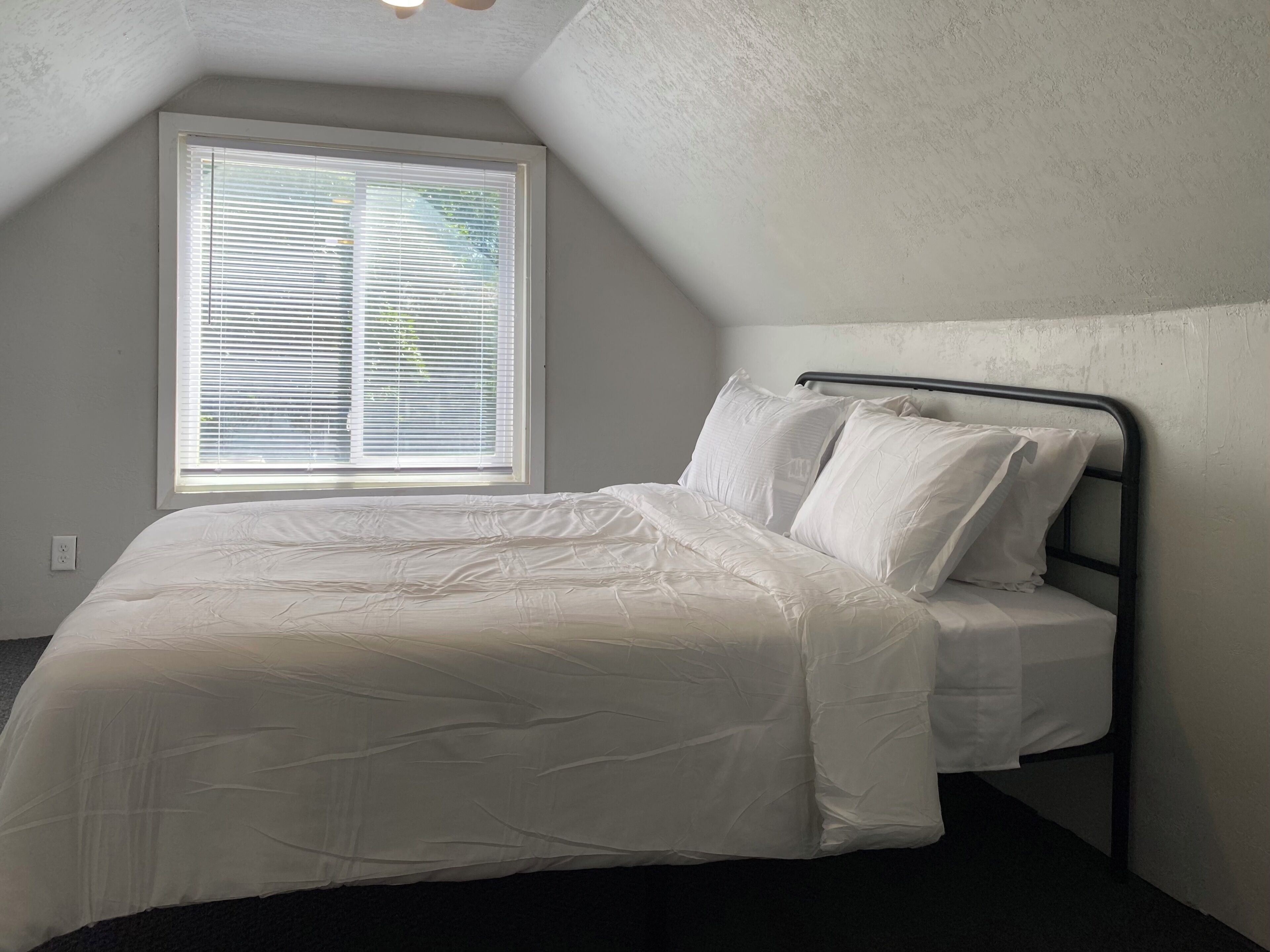 Een eenpersoonsbed met metalen frame in een kamer met schuine wand en raam met zonwering.