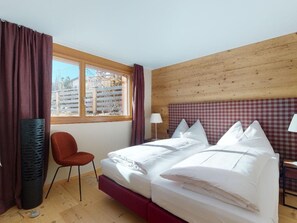 A hotel room with a wooden headboard, a bed with white linens, a red chair, a window with curtains, and a view of the outside. - Apartment Quadrellas 4.5 by Interhome (Samedan)