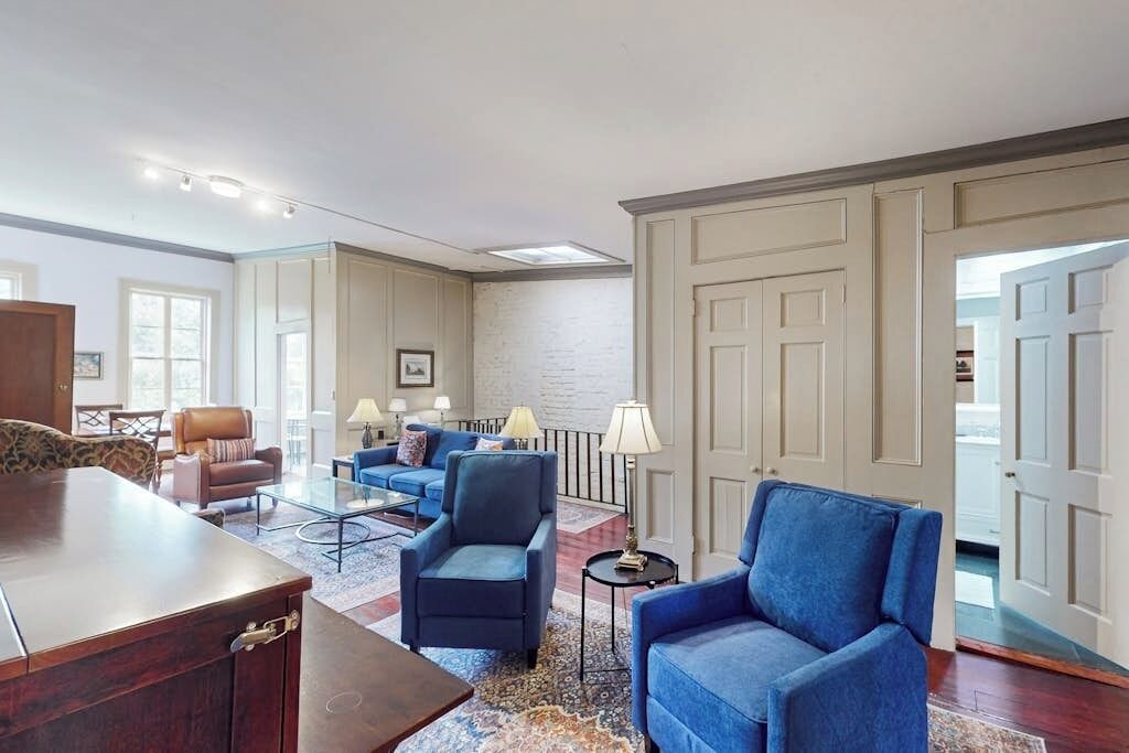 Een ruime woonkamer met een groot houten bureau, diverse zitplaatsen waaronder een blauwe bank en fauteuils, en een kamerbreed tapijt met dessin.