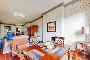 Un coin repas avec une table en bois, des chaises et un saladier. Il y a un mur en briques blanches et un tableau accroché au mur.