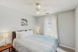Ein Schlafzimmer mit einem Bett, einem Nachttisch, einem Deckenventilator, einem Fenster mit Vorhängen und einem an der Wand hängenden Kunstwerk.