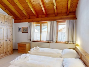 Ein Zimmer mit Holzbalken, einem Bett, einem Nachttisch, einer Kommode und einem Fenster mit Vorhängen.
