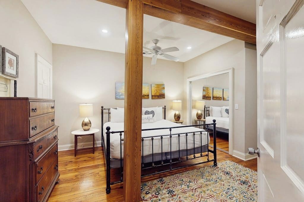 A bedroom with a bed, wooden floor, ceiling fan, and a large mirror.