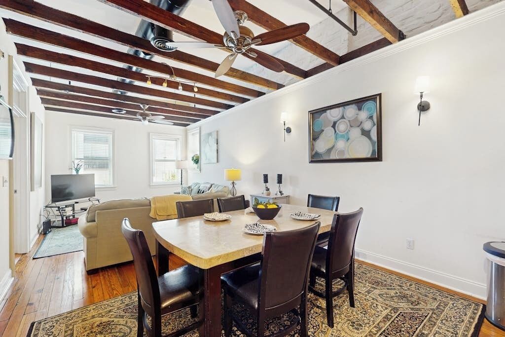 Uma sala de jantar com mesa e cadeiras de madeira, um ventilador de teto e um quadro na parede.